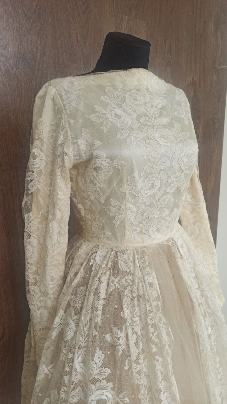 Op de afbeelding: Ivoor kanten trouwjurk met lange mouwen en een hoge halslijn. De jurk heeft ingewikkelde bloemenkanten details over de hele bodice en rok, waardoor een romantische en elegante look ontstaat. De rok heeft een vloeiend, gelaagd ontwerp.
