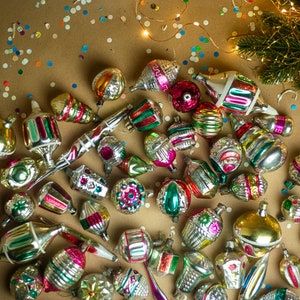 May include: A collection of vintage glass Christmas ornaments in various colors and designs, including silver, gold, red, green, and blue. The ornaments are scattered on a brown surface with confetti.