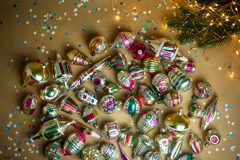May include: A collection of vintage glass Christmas ornaments in various colors and designs, including silver, gold, red, green, and blue. The ornaments are scattered on a brown surface with confetti.