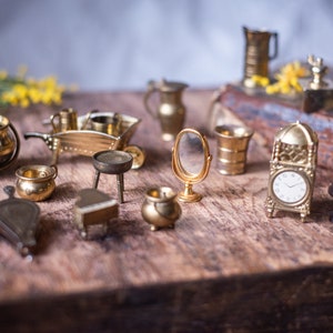 Op de afbeelding: Een verzameling miniatuurvoorwerpen van verguld metaal, waaronder een klok, een spiegel, een kruiwagen, een ketel en een kruk. De voorwerpen zijn gerangschikt op een houten oppervlak.