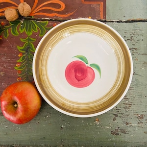 May include: A white ceramic bowl with a hand-painted red peach design and green leaves. The bowl has a brown and gold rim. An apple and walnuts are next to the bowl on a distressed wooden surface.