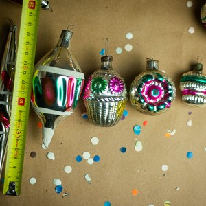 May include: A collection of vintage glass Christmas ornaments with intricate designs and colorful details. The ornaments are silver with green, pink, and blue accents. The ornaments are arranged on a brown surface with confetti.