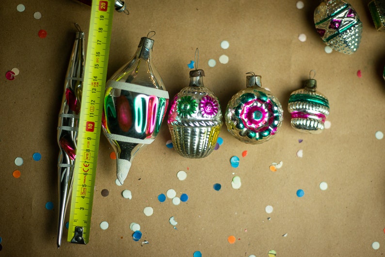 May include: A collection of vintage glass Christmas ornaments with intricate designs and colorful details. The ornaments are silver with green, pink, and blue accents. The ornaments are arranged on a brown surface with confetti.