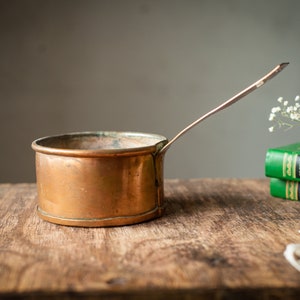 Pode incluir: Uma panela de cobre com uma alça longa, colocada sobre uma superfície de madeira. A panela tem um corpo redondo e um acabamento ligeiramente manchado. Flores secas e livros verdes ao fundo.