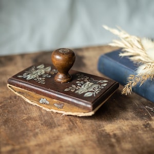 May include: A vintage wooden inkwell with a floral design and a wooden lid. The inkwell is sitting on a wooden surface.