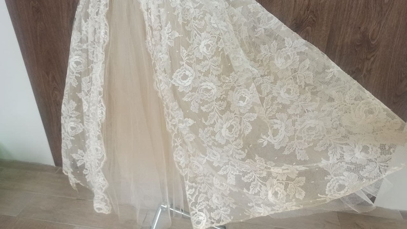 Op de afbeelding: Ivoor kanten stof met een bloemmotief, gedrapeerd over lichtbeige tule. Het kant heeft gedetailleerde rozen- en bladerontwerpen. De stof wordt waarschijnlijk gebruikt voor bruids- of formele kleding.