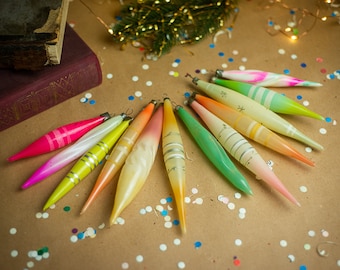 May include: A collection of twelve elongated, tapered glass ornaments arranged in a fan shape. Each ornament features a unique color gradient, including shades of pink, yellow, orange, green, and white, with delicate painted line details.