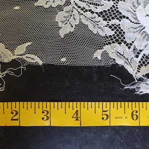 Op de afbeelding: Close-up van ivoorkleurige kanten stof met bloempatronen. Het kant wordt gemeten met een gele meetlint, met ongeveer 20 cm van de stof. Het kant heeft een delicate, open weefsel en wordt waarschijnlijk gebruikt voor kleding of knutselprojecten.