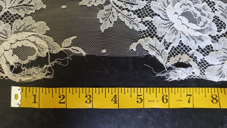 Op de afbeelding: Close-up van ivoorkleurige kanten stof met bloempatronen. Het kant wordt gemeten met een gele meetlint, met ongeveer 20 cm van de stof. Het kant heeft een delicate, open weefsel en wordt waarschijnlijk gebruikt voor kleding of knutselprojecten.