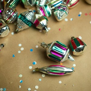 May include: A collection of vintage Christmas ornaments in various colors and designs, including silver, green, red, and pink, scattered on a brown paper background with white and blue confetti.