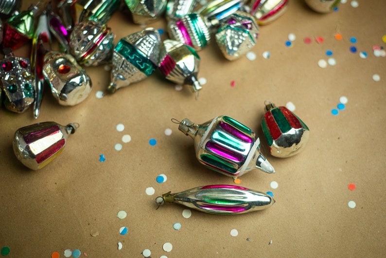 May include: A collection of vintage Christmas ornaments in various colors and designs, including silver, green, red, and pink, scattered on a brown paper background with white and blue confetti.