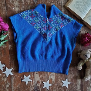 Op de afbeelding: Een blauwe mouwloze trui met een geborduurde halslijn. De trui heeft een rits en een geribde zoom. De afbeelding bevat ook een boeket roze pioenrozen, een teddybeer, een boek en een sterrenslingers op een houten oppervlak.