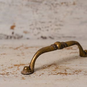 May include: A vintage-style brass drawer pull with a curved handle and decorative detailing. The hardware has an aged, antique finish, suggesting it is for furniture restoration or a rustic design aesthetic.
