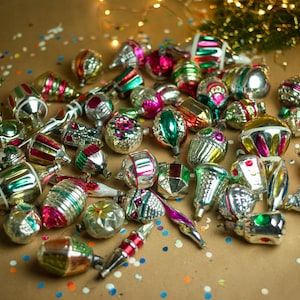 May include: A collection of vintage glass Christmas ornaments in various colors and designs, including silver, gold, red, green, and blue. The ornaments are scattered on a brown surface with confetti.