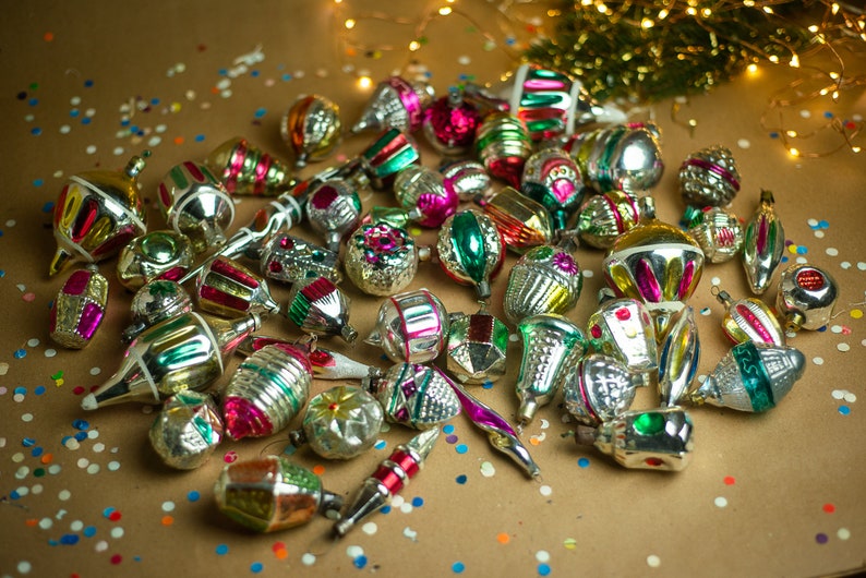 May include: A collection of vintage glass Christmas ornaments in various colors and designs, including silver, gold, red, green, and blue. The ornaments are scattered on a brown surface with confetti.