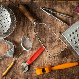 May include: A collection of vintage kitchen tools, including a potato masher, grater, can opener, and nutcracker, arranged on a rustic wooden surface. The tools are made of metal and wood, with some featuring red and orange handles.