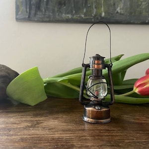 May include: A miniature metal lantern featuring a copper-toned finish, a clear glass globe, and a thin black wire handle. The lantern has a circular base, a central burner housing, and a protective wire cage surrounding the glass component.