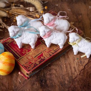 May include: Five white ceramic sheep ornaments, each featuring a small satin ribbon tied around its neck in light blue, pink, or yellow. The sheep are arranged on top of a vintage red and gold book, accompanied by one blue and one orange egg.
