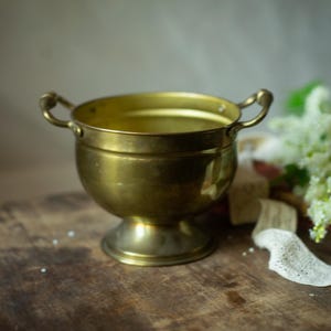 Pode incluir: Uma taça de latão polido com corpo arredondado, base de pedestal e duas pequenas alças curvas nos lados opostos. A superfície metálica tem um tom dourado quente com variações subtis de brilho e um acabamento liso e polido.