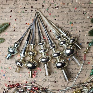 Può includere: Una collezione di cinque puntali per albero in vetro color argento, ciascuno caratterizzato da una lunga guglia appuntita e una base bulbosa. Gli ornamenti hanno una finitura metallica riflettente e sono disposti a ventaglio con fili di perline.