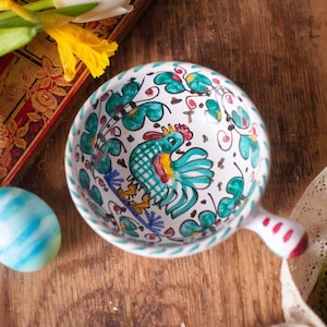 May include: A white ceramic bowl with a handle, featuring a hand-painted green and blue rooster motif in the center, surrounded by decorative green floral patterns and small red accents along the rim and handle, with a smooth, glossy finish.
