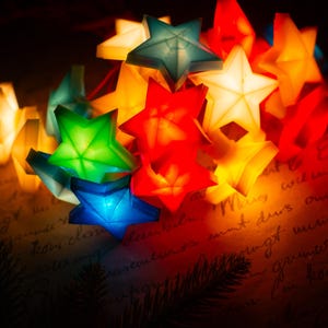 Puede incluir: Una cadena de luces con forma de estrella en tonos vibrantes de rojo, azul, verde y amarillo. Cada estrella tiene una superficie translúcida con líneas geométricas internas visibles, proyectando un brillo cálido sobre un texto manuscrito.