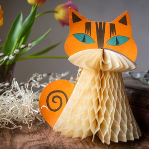 May include: An orange paper cat decoration with a honeycomb body and a heart-shaped tail. The cat has teal eyes and black stripes. A vase of yellow tulips and pussy willow branches are in the background.