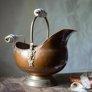 May include: A copper coal scuttle with a brass base and a brass handle featuring a lion head mount. The handle and the side grip are accented with white and blue floral porcelain inserts. The vessel has a wide, curved spout and a polished finish.