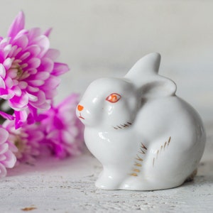 Op de afbeelding: Een wit keramieken konijnbeeldje met oranje ogen en gouden accenten, naast levendige roze en witte chrysantenbloemen. Het konijn is een decoratief item, mogelijk voor Pasen of de lente.