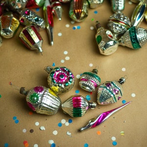 May include: A collection of vintage glass Christmas ornaments with silver, green, pink, and gold accents. The ornaments are scattered on a brown paper background with colorful confetti.