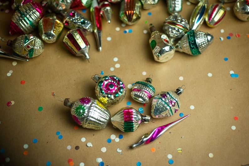 May include: A collection of vintage glass Christmas ornaments with silver, green, pink, and gold accents. The ornaments are scattered on a brown paper background with colorful confetti.