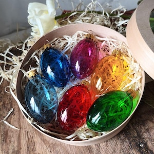 Może przedstawiać: Okrągłe drewniane pudełko wypełnione kolorowymi ozdobami wielkanocnymi w kształcie jajek: niebieskim, fioletowym, pomarańczowym, zielonym i czerwonym. Każde jajko ma złotą nasadkę i jest wypełnione teksturowanym materiałem. Jajka są ułożone w słomie.