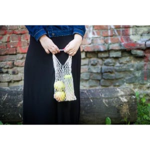 May include: A white string mesh bag containing two round yellow and green apples and a tall cylindrical bottle with a light green cap. The bag is held by two hands, with the fingers gripping the top edge of the woven netting material.