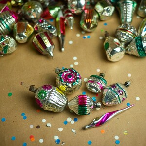 May include: A collection of vintage glass Christmas ornaments with silver and gold paint, featuring colorful designs and patterns. The ornaments are scattered on a brown paper background with confetti.