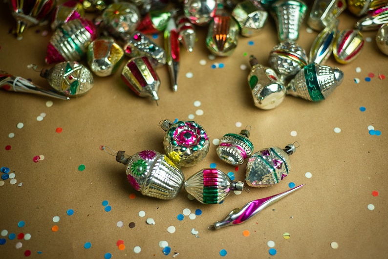 May include: A collection of vintage glass Christmas ornaments with silver and gold paint, featuring colorful designs and patterns. The ornaments are scattered on a brown paper background with confetti.
