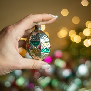 May include: A vintage glass Christmas ornament with a silver finish and geometric green, red, and yellow patterns. The ornament is being held by a hand against a blurred background of lights.