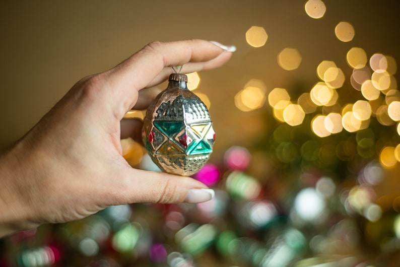 May include: A vintage glass Christmas ornament with a silver finish and geometric green, red, and yellow patterns. The ornament is being held by a hand against a blurred background of lights.
