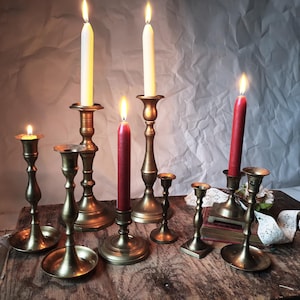 Könnte beinhalten: Eine Sammlung antiker Messing-Kerzenhalter mit brennenden Kerzen. Die Kerzen sind weiß und rot, und die Halter variieren in Größe und Design. Die Anordnung befindet sich auf einer rustikalen Holzoberfläche und erzeugt eine warme, Vintage-Ästhetik.