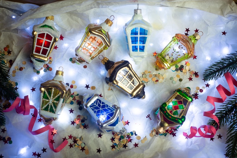 May include: Assortment of eight miniature glass lantern Christmas ornaments.  Styles include red, green, blue, and gold with varied window pane designs.  Gold-colored metal accents are visible on several.  Ornaments are displayed on a white surface with pink ribbon and confetti.