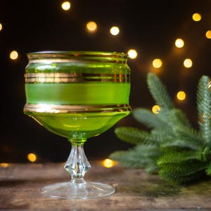 May include: A vintage green glass goblet with gold trim. The goblet has a wide bowl and a clear, faceted stem. The glass is filled with a green liquid. The background is dark with blurred lights and a sprig of evergreen.