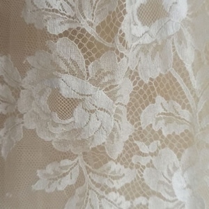 Op de afbeelding: Close-up van witte bloemenkanten stof. Het delicate kant heeft een herhalend patroon van bloemen en bladeren, waardoor een textuurrijk en elegant ontwerp ontstaat. De achtergrond is een zachte, neutrale kleur, die de ingewikkelde details van het kant benadrukt.