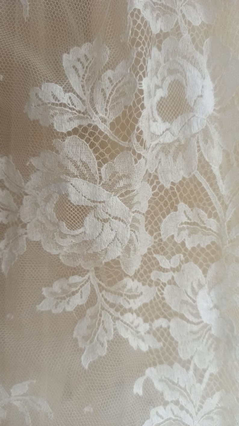 Op de afbeelding: Close-up van witte bloemenkanten stof. Het delicate kant heeft een herhalend patroon van bloemen en bladeren, waardoor een textuurrijk en elegant ontwerp ontstaat. De achtergrond is een zachte, neutrale kleur, die de ingewikkelde details van het kant benadrukt.