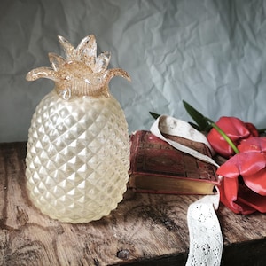 May include: A clear glass pineapple figurine with a textured diamond-patterned body and a gold-flecked crown. The pineapple is positioned next to a vintage red book with gold-edged pages and a cluster of three vibrant red tulip flowers.