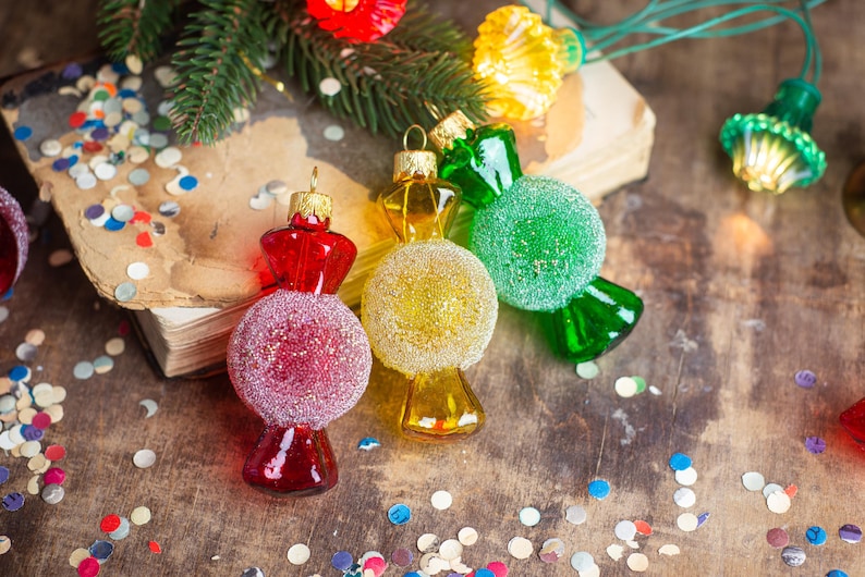 May include: Four candy cane shaped Christmas ornaments, one red, one green, one yellow, and one clear with glitter. The ornaments are on a wooden surface with confetti.