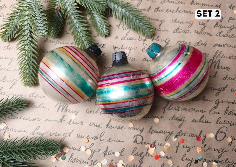 Puede incluir: Tres adornos navide&ntilde;os esf&eacute;ricos de vidrio con rayas horizontales en tonos azul verdoso, rosa y dorado sobre una base plateada. Cada adorno tiene una tapa met&aacute;lica en la parte superior. El texto SET 2 es visible en la esquina superior derecha.