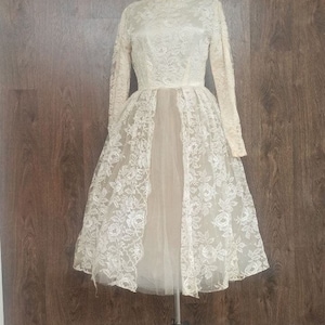 Op de afbeelding: Ivoor vintage jurk met lange mouwen en een hoge halslijn. De jurk heeft een delicate bloemen kanten overlay op het lijfje en de rok, met een doorschijnende, vloeiende onderlaag. De jurk wordt getoond op een metalen paspop tegen een achtergrond van houten panelen.