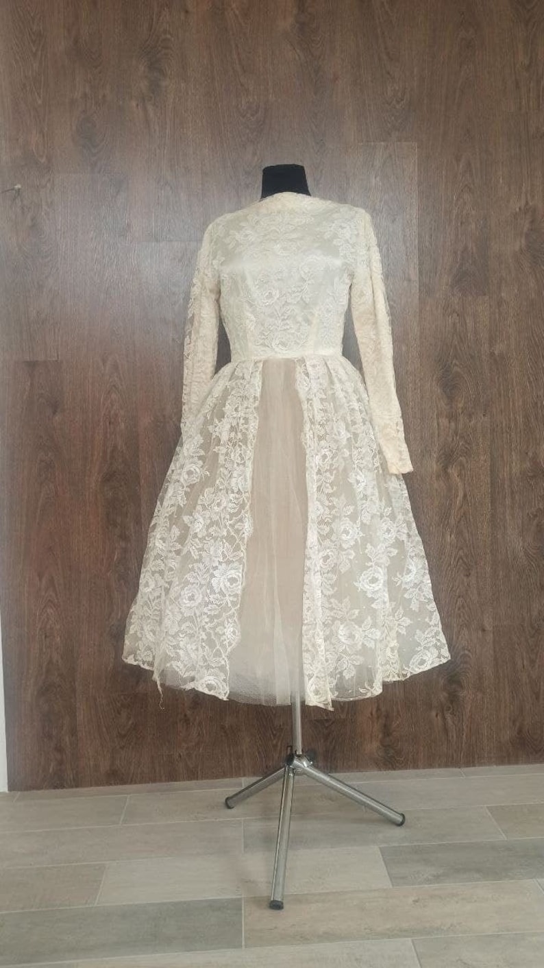 Op de afbeelding: Ivoor vintage jurk met lange mouwen en een hoge halslijn. De jurk heeft een delicate bloemen kanten overlay op het lijfje en de rok, met een doorschijnende, vloeiende onderlaag. De jurk wordt getoond op een metalen paspop tegen een achtergrond van houten panelen.