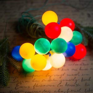 Puede incluir: Una cadena de luces LED esféricas que presenta una variedad de colores translúcidos, incluyendo rojo, amarillo, verde, azul y blanco. Las bombillas redondas están conectadas por un cable fino y dispuestas en un grupo, creando un brillo multicolor.