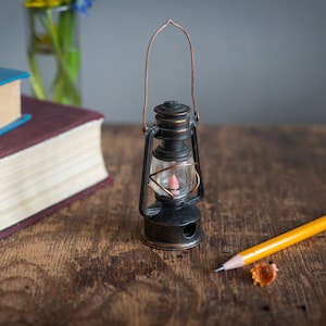 May include: A miniature black lantern with a copper handle and a pink light sits on a wooden surface. A yellow pencil and two books are also on the table. The books have blue and burgundy covers.