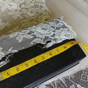 Op de afbeelding: Ivoor kanten stof met bloempatronen, een zwarte doos en een gele meetlint. De tape geeft een meting van ongeveer 23 cm aan. Het kant is over de doos gedrapeerd.